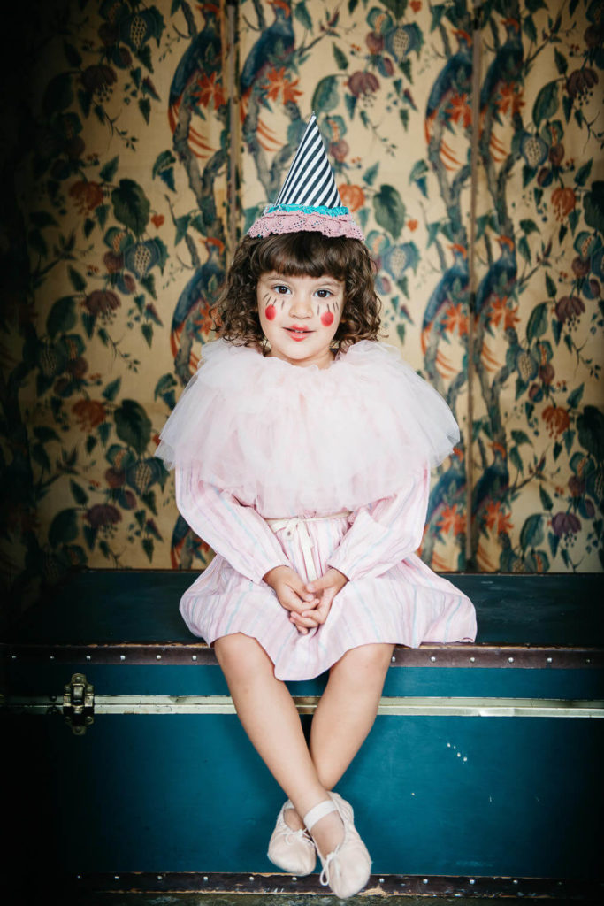 A young child with curly hair, dressed in a striped clown outfit and hat, sits on a vintage trunk. The child has painted cheeks and a ruffled collar. The background features a floral-patterned wallpaper with birds.
