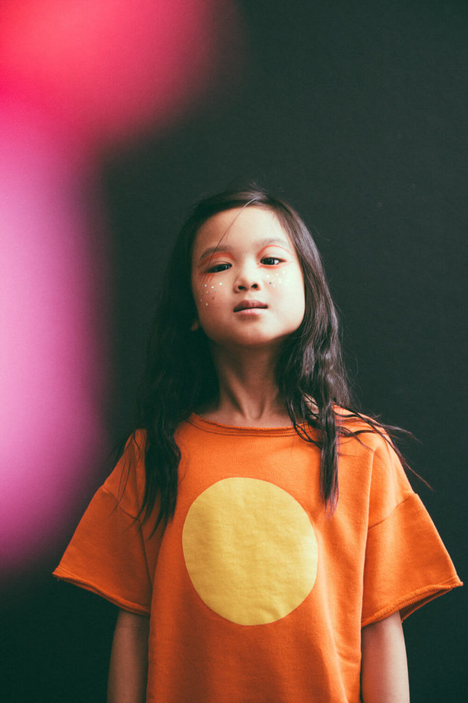 A young girl with long dark hair stands against a black background, wearing an orange shirt with a large yellow circle. Her face is adorned with small star-shaped stickers, and there is a blurred pink shape in the foreground.