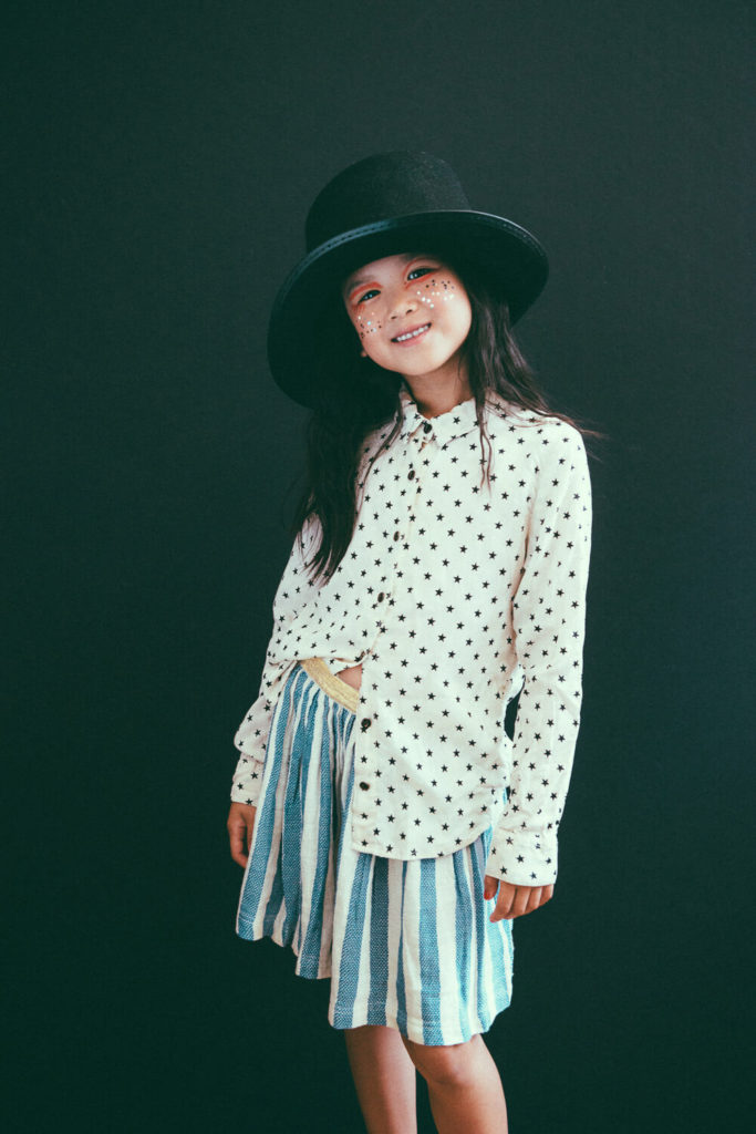 A young girl smiles against a dark background. She wears a black hat, a white blouse with star patterns, and a blue and white striped skirt. Her cheeks are decorated with colorful stickers.