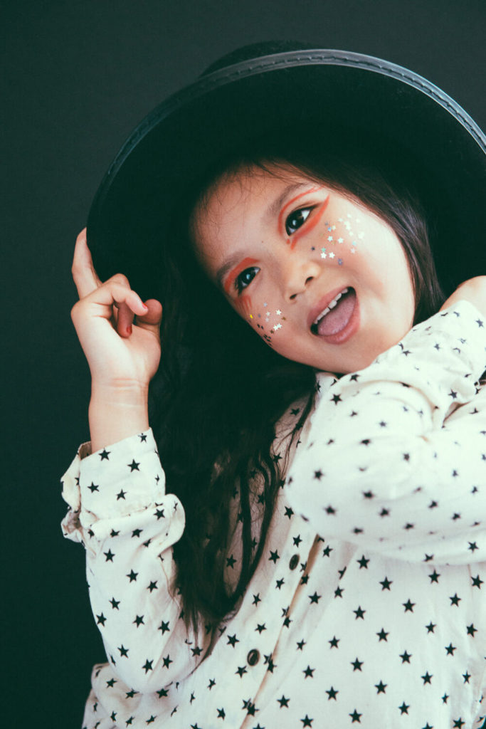 A young child wears a black hat and a white shirt with black stars. They have star-shaped stickers on their cheeks and orange makeup around their eyes. The child is smiling playfully against a dark background.