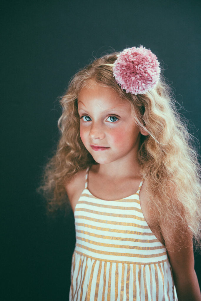 A young girl with long, curly blonde hair stands against a dark background. She wears a striped dress and has a large pink pom-pom hair accessory. She looks slightly to the side with a gentle expression.