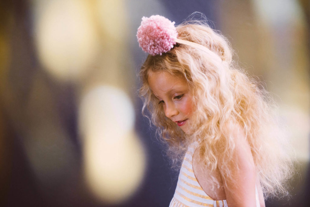 A young girl with long curly hair and a pink pom-pom headband looks down, smiling slightly. She is wearing a striped dress, and the background is softly blurred with warm, bokeh lighting.