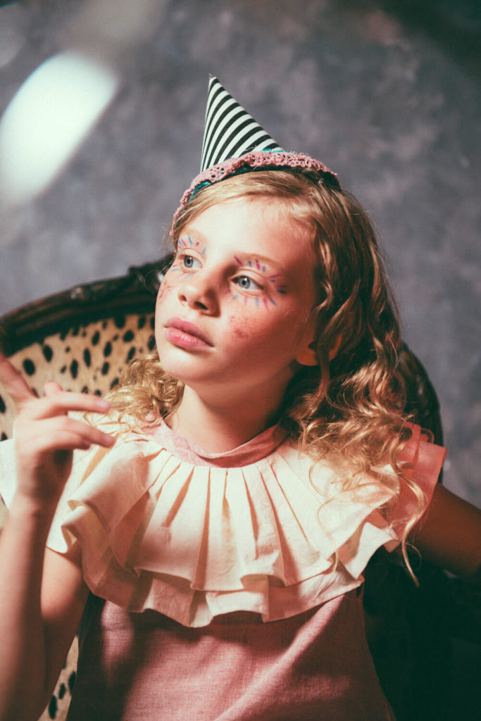 A child with curly hair wears a party hat and a frilly collar. They are sitting in a chair, gazing upward with a thoughtful expression, painted makeup patterns on their face. The background is softly lit and colorful.
