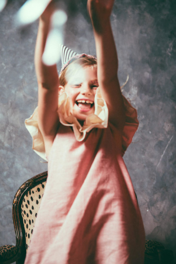 A joyful child wearing a pink dress and a striped party hat, raising their arms in excitement. They sit on a vintage chair against a textured background, smiling happily.