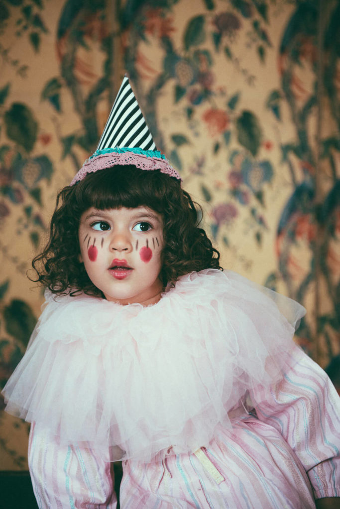 A child dressed as a clown wears a striped party hat and has face paint with red heart designs on cheeks. They have curly hair and are wearing a pink ruffled costume. The background features a floral patterned wallpaper.