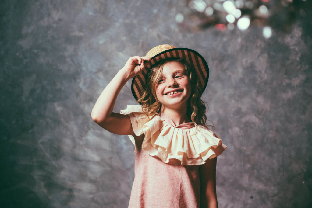 A smiling young girl with curly hair wears a wide-brimmed hat and a ruffled dress, posing against a textured gray background. She holds the brim of the hat with her hand, and her expression is joyful and playful.