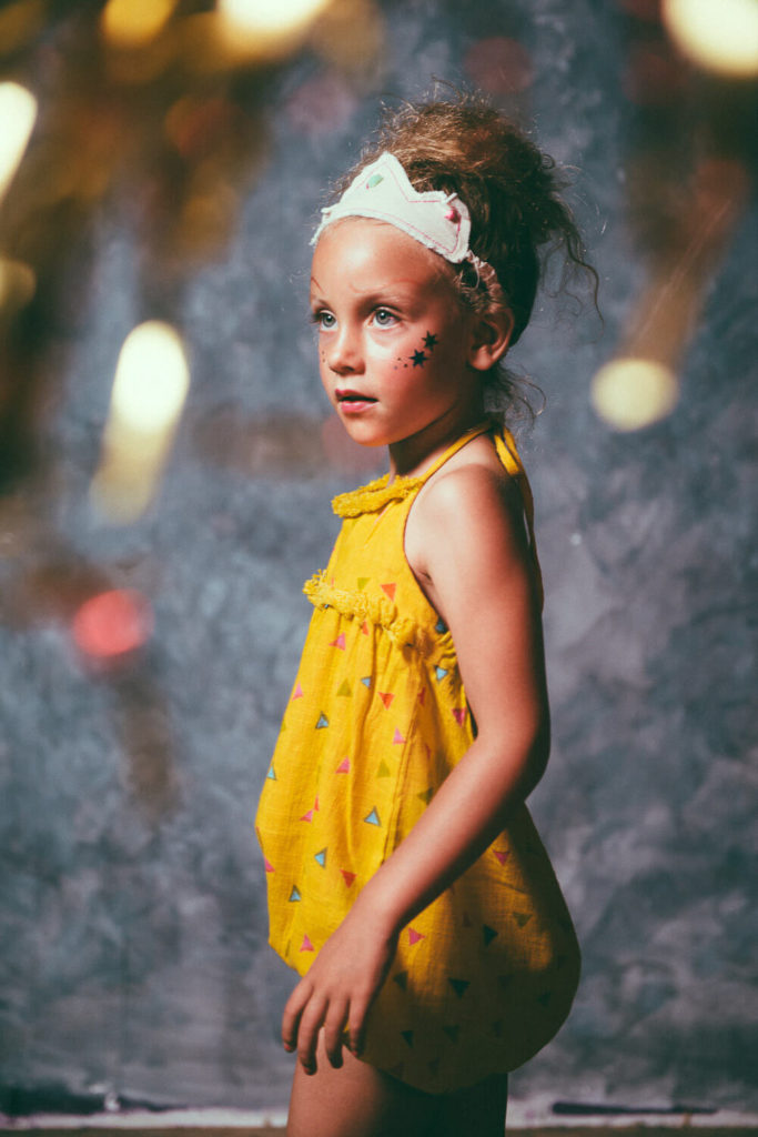 A young girl with curly hair stands sideways, wearing a yellow dress with colorful shapes. She has painted stars on her cheek and a decorative headband. The background is a blurred mix of gray and golden hues.