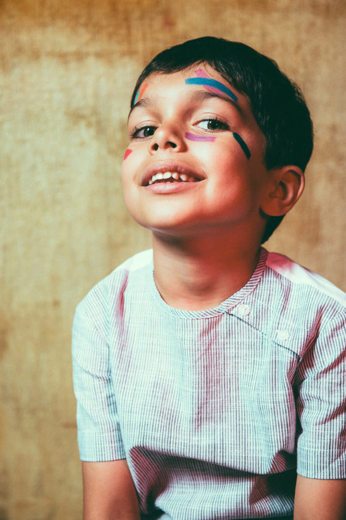 A young boy with short dark hair smiles with colorful paint streaks on his face. He is wearing a light blue and white striped shirt and is posed against a brown textured background.