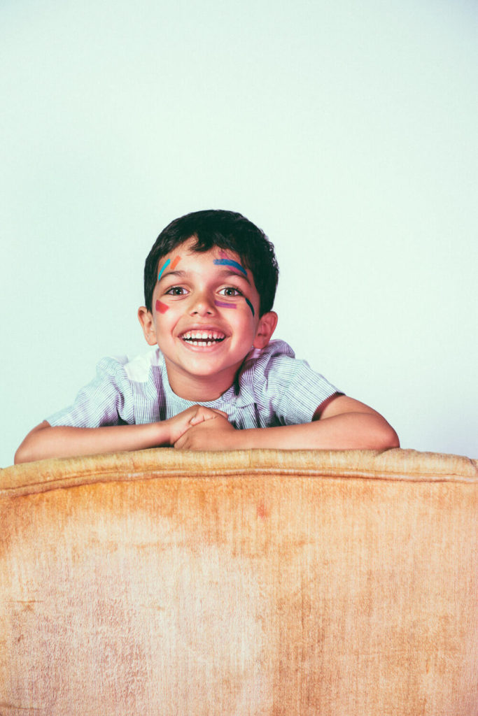 A smiling child leans on the back of a beige sofa. The child has colorful face paint with stars on their cheeks. They wear a striped shirt and have short dark hair, set against a plain light background.