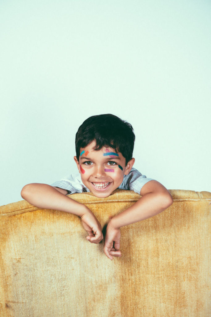 A smiling child with face paint leans on the back of a beige sofa, looking directly at the camera.