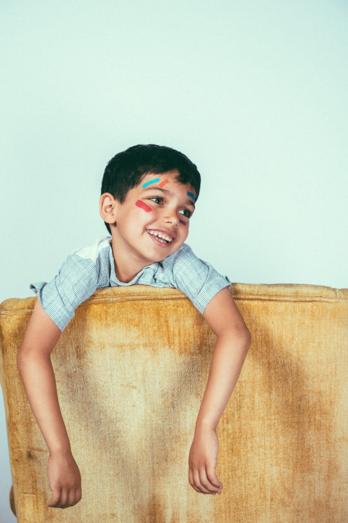 A young child with colorful face paint smiles joyfully while leaning over the back of a yellow sofa. He wears a blue checkered shirt and looks happy against a plain background.