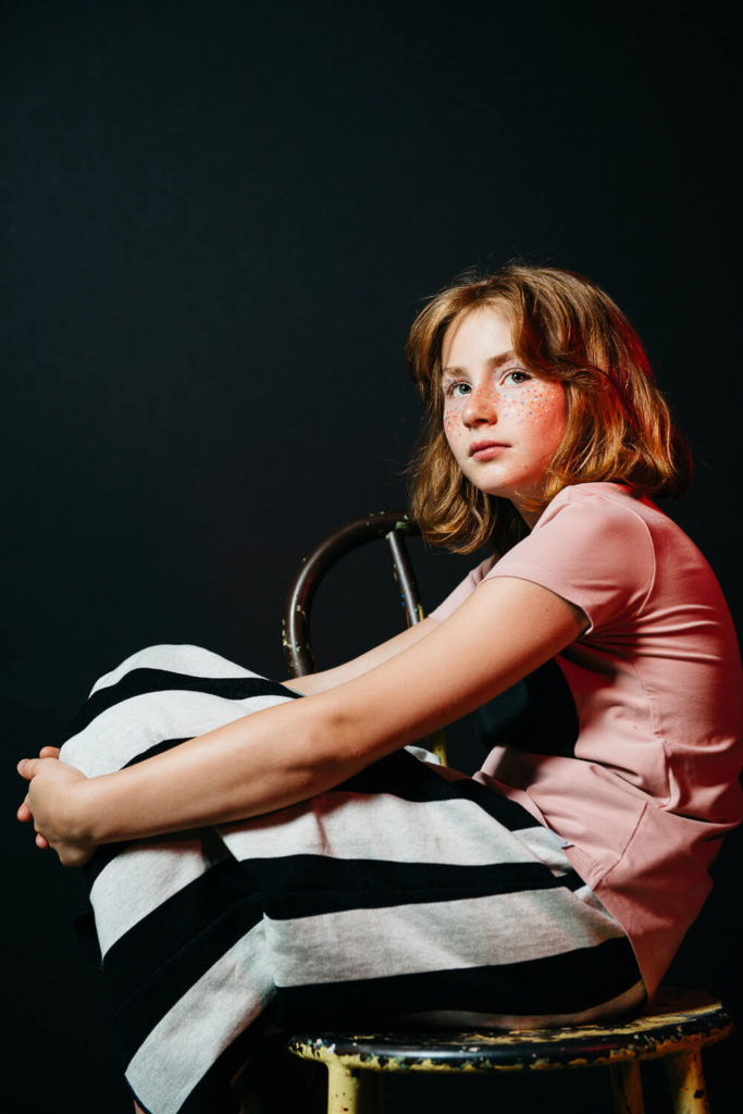 A young girl with shoulder-length hair and freckles sits on a stool against a dark background. She wears a pink shirt and a black-and-white striped skirt, gazing thoughtfully to the side.