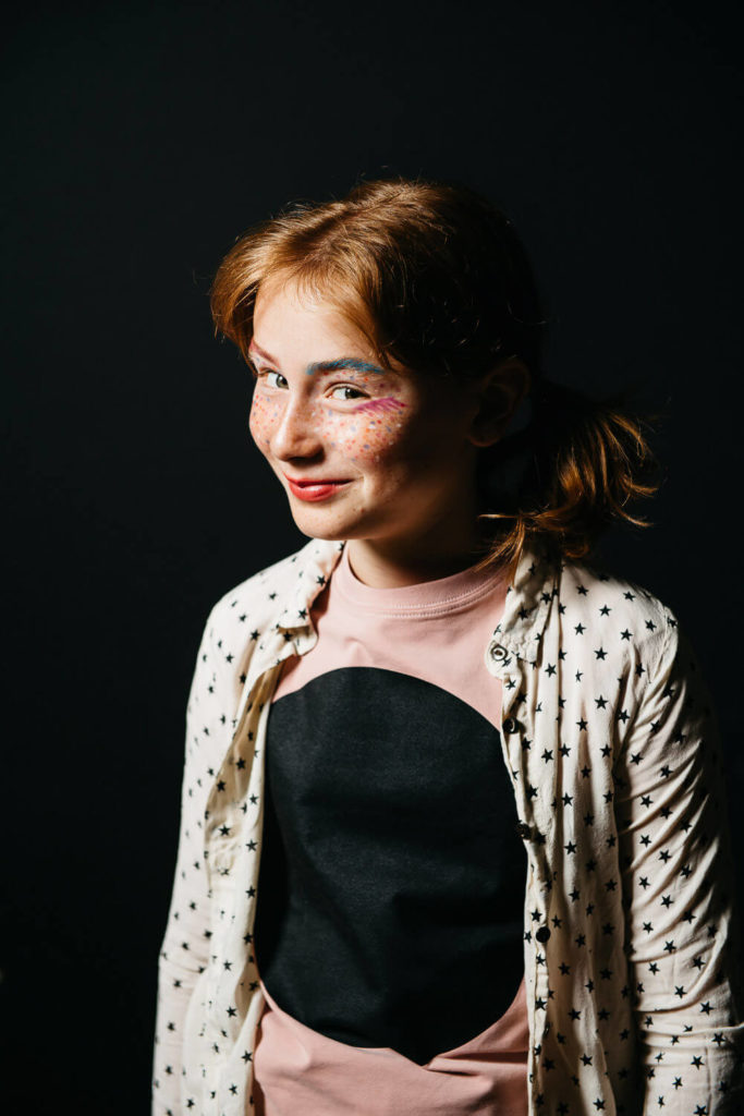 A young girl with red hair smiles while wearing a star-patterned shirt over a pink top. Her face is adorned with colorful makeup, including pink eyeshadow and white polka dots. The background is dark.