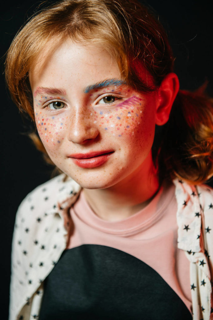 A young person with freckles and colorful makeup smiles gently. They have red hair and wear a light shirt with star patterns over a pink top. The background is dark, with a red light casting a glow on one side of their face.