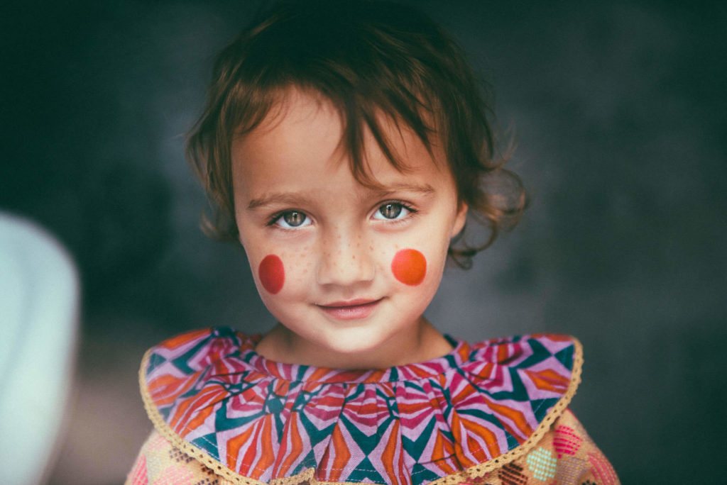 A child with green eyes and rosy cheeks smiles at the camera. They are wearing a colorful outfit with bold patterns and have two red circles painted on their cheeks. The background is softly blurred.