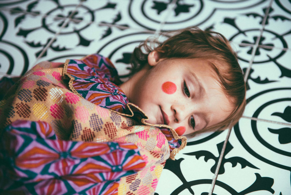 A young child with red face paint on their cheeks is wrapped in a colorful patterned fabric, lying on a black and white patterned tile floor. The child has a thoughtful expression.