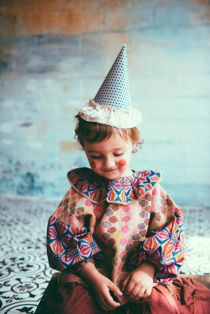 A young child dressed as a clown with a colorful costume, frilled collar, and polka-dotted cone hat sits on a patterned floor. The child smiles adorably with rosy cheeks and closed eyes, surrounded by a soft, textured background in blue and gold.