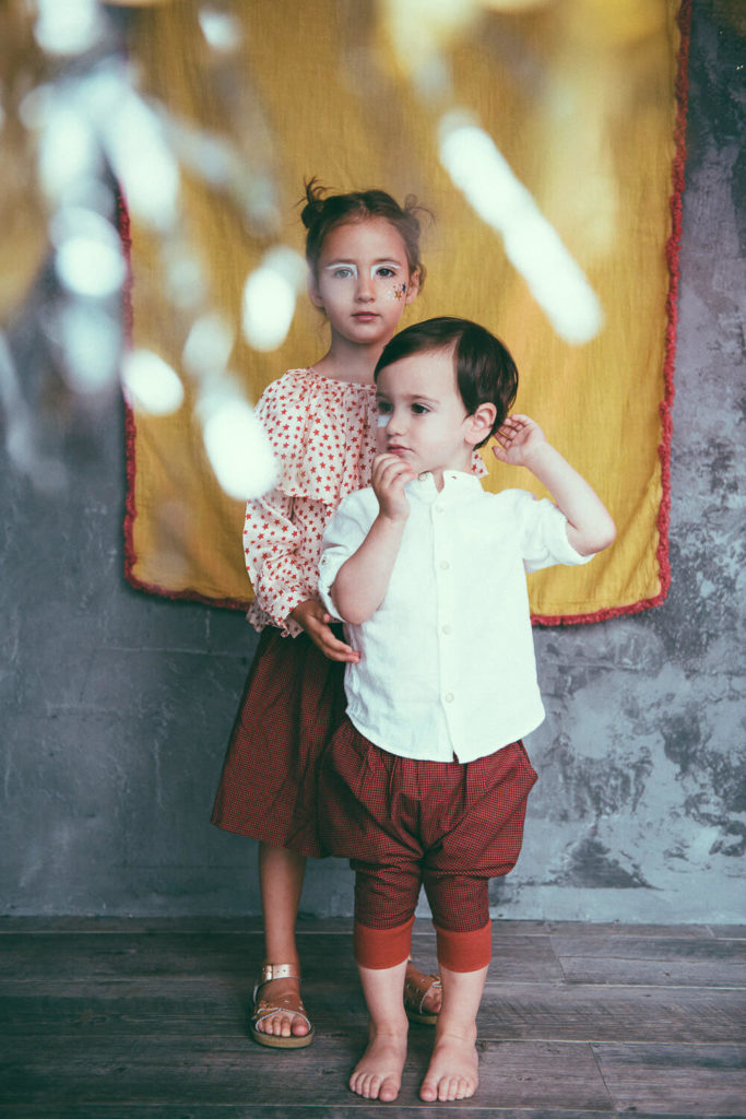 A young girl and a toddler stand together indoors. The girl is wearing a polka dot blouse and red skirt, while the toddler wears a white shirt and red shorts. They are in front of a textured gray wall with a yellow cloth hanging behind them.