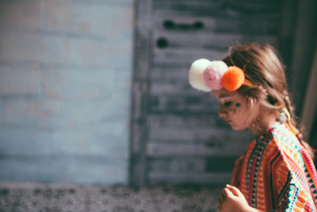 A child dressed in colorful attire stands in profile against a blurred rustic background. They have poms in their hair and appear thoughtful or focused.