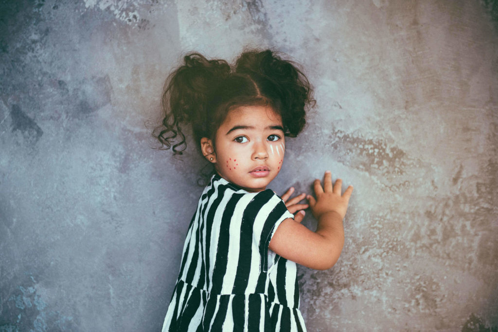 A young girl with curly hair and face paint, wearing a black and white striped dress, stands against a textured wall. She looks over her shoulder with a curious expression.