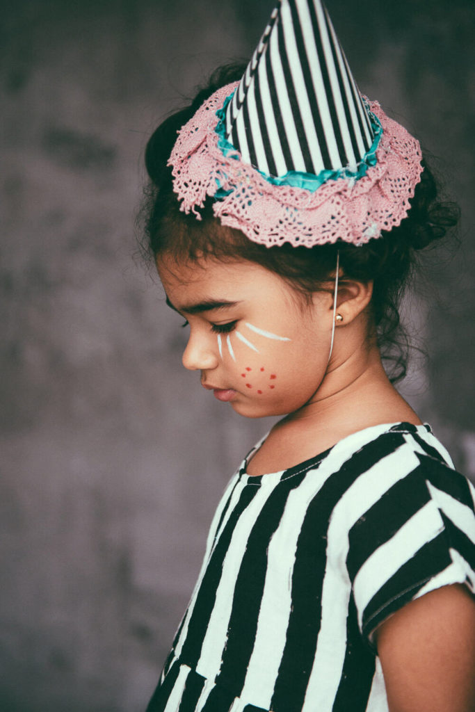 A young girl wearing a striped outfit and matching hat with lace details stands in profile. Her face is painted with red dots and white lines, resembling a clown. The background is blurred and gray, adding a soft contrast to her focused expression.