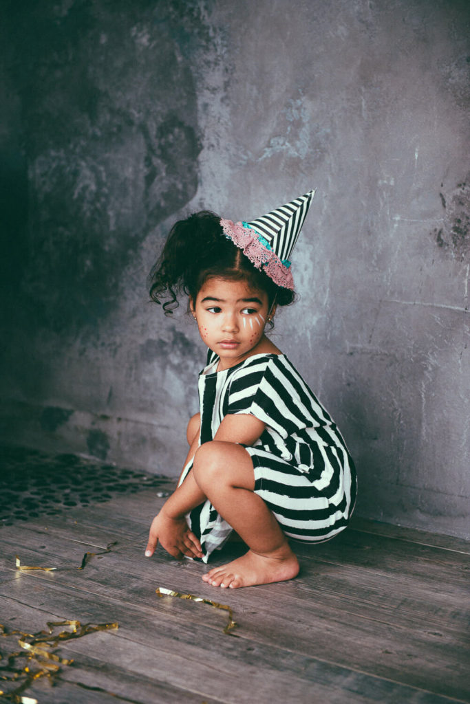 A young girl wearing a black and white striped dress and a whimsical party hat sits against a textured gray wall. She is barefoot with a curious expression, surrounded by scattered gold ribbon on the wooden floor.