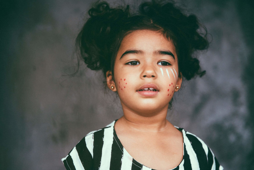 A young girl with curly hair and painted tribal designs on her face, wearing a black and white striped top, stands against a dark background.