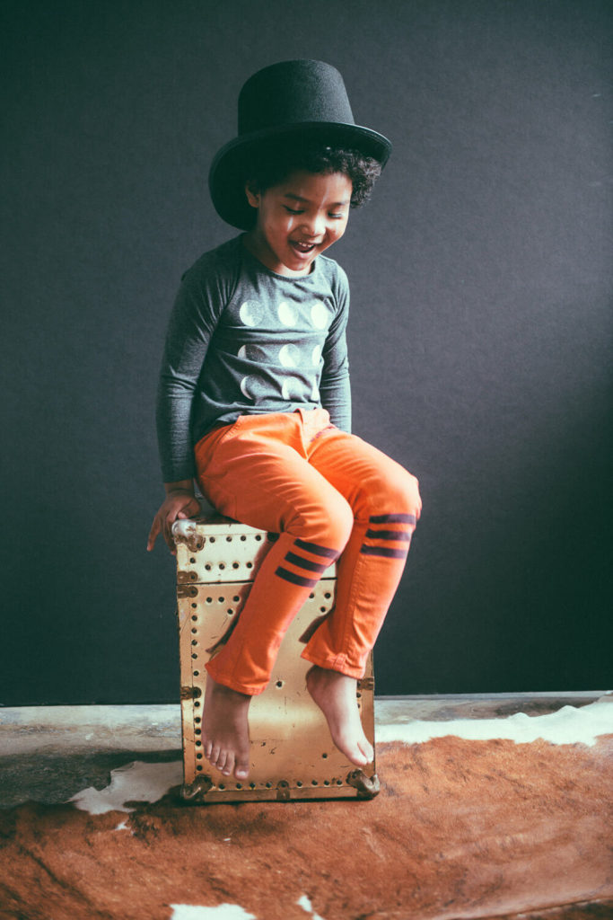 A smiling child wearing a top hat sits playfully on a box. They are dressed in a gray shirt with a pattern and orange pants with stripes. The backdrop is dark, and the floor has a cowhide rug.