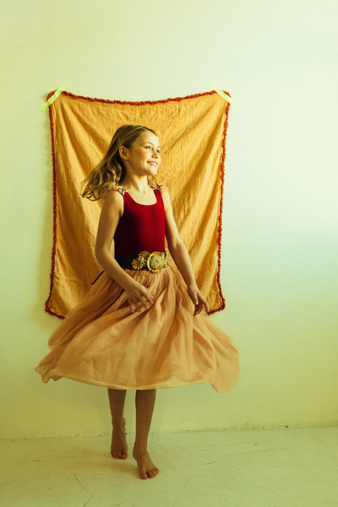 A girl with long hair twirls barefoot in a flowing peach skirt and red top. She stands in front of a mustard-colored textured backdrop, smiling, with her skirt fanned out around her. The setting is warmly lit.
