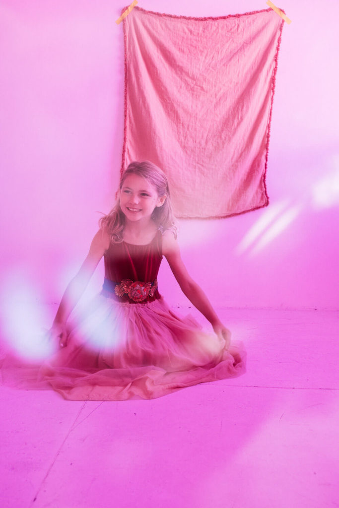A young girl in a pink tulle dress with a floral belt sits on the floor in a room with pink lighting. Behind her, a pink fabric is taped to the wall. Gentle beams of light create a soft, dreamy atmosphere.