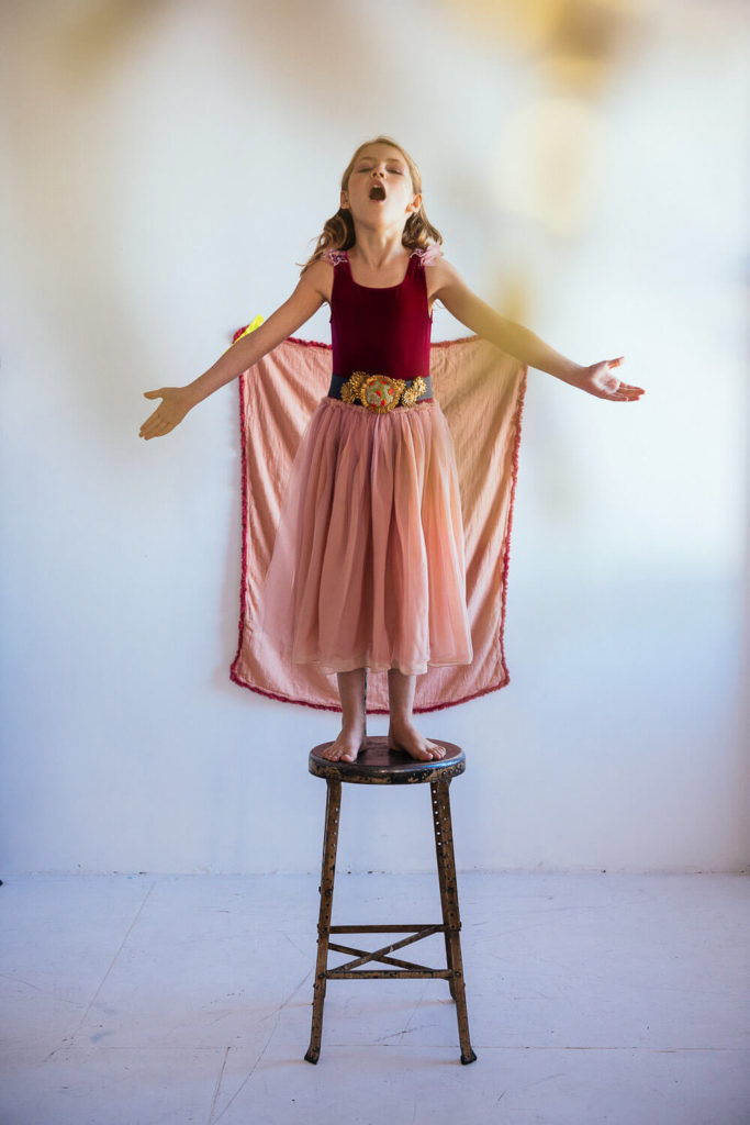 A girl stands barefoot on a wooden stool, arms outstretched, eyes closed. She wears a pink tulle skirt and a burgundy top with a decorative belt. A pink garment is draped behind her, resembling wings. The setting is a softly lit room.