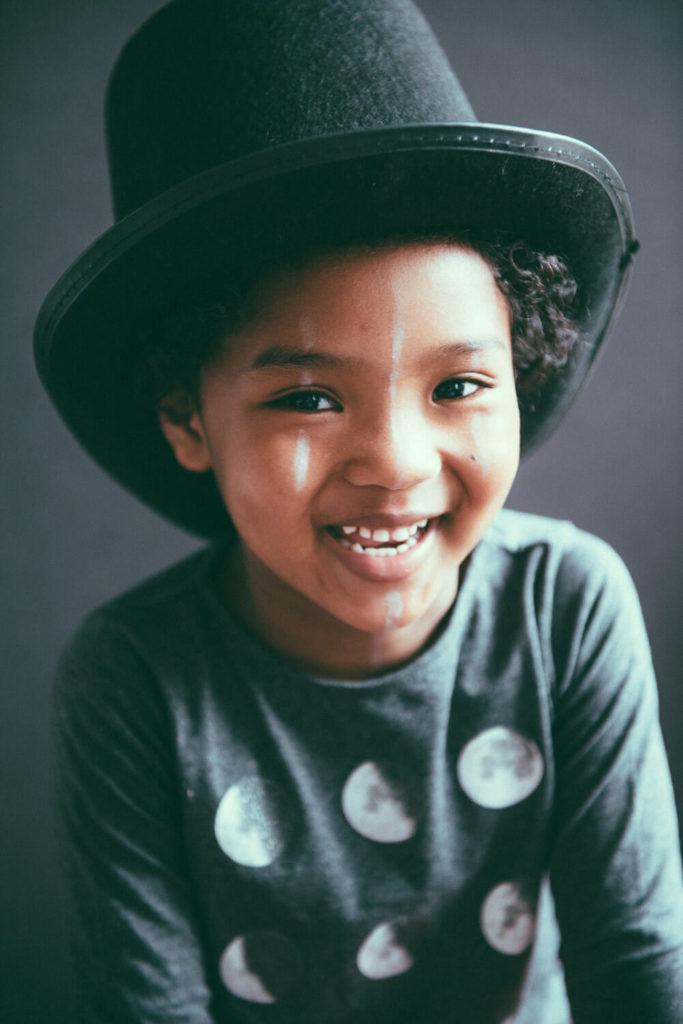 A smiling child wearing a large top hat and a shirt with moon phases sits against a dark background.