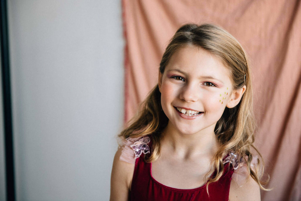 A smiling young girl with long blonde hair stands in front of a pink curtain. She is wearing a red dress with lace details on the shoulders and has star-shaped stickers on her cheek.