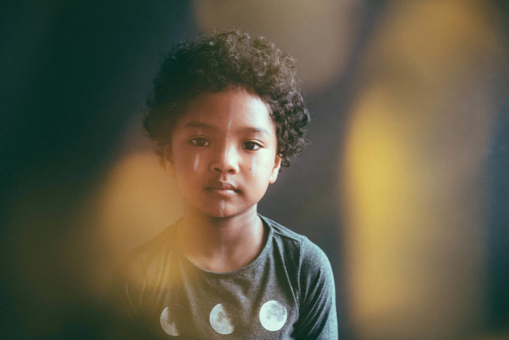 A young child with curly hair looks forward with a calm expression. They are wearing a shirt with moon phases printed on it. The background is softly blurred with warm light bokeh effects.