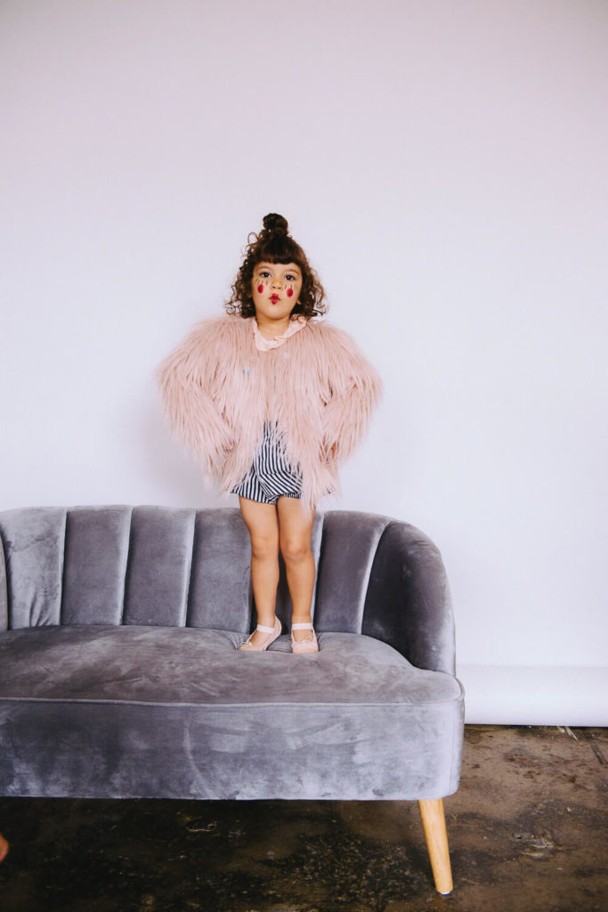 A young child stands on a gray velvet couch wearing a fluffy pink coat and striped outfit. They have curly hair styled in a top knot, round glasses, and pink shoes, posing confidently against a plain white background.