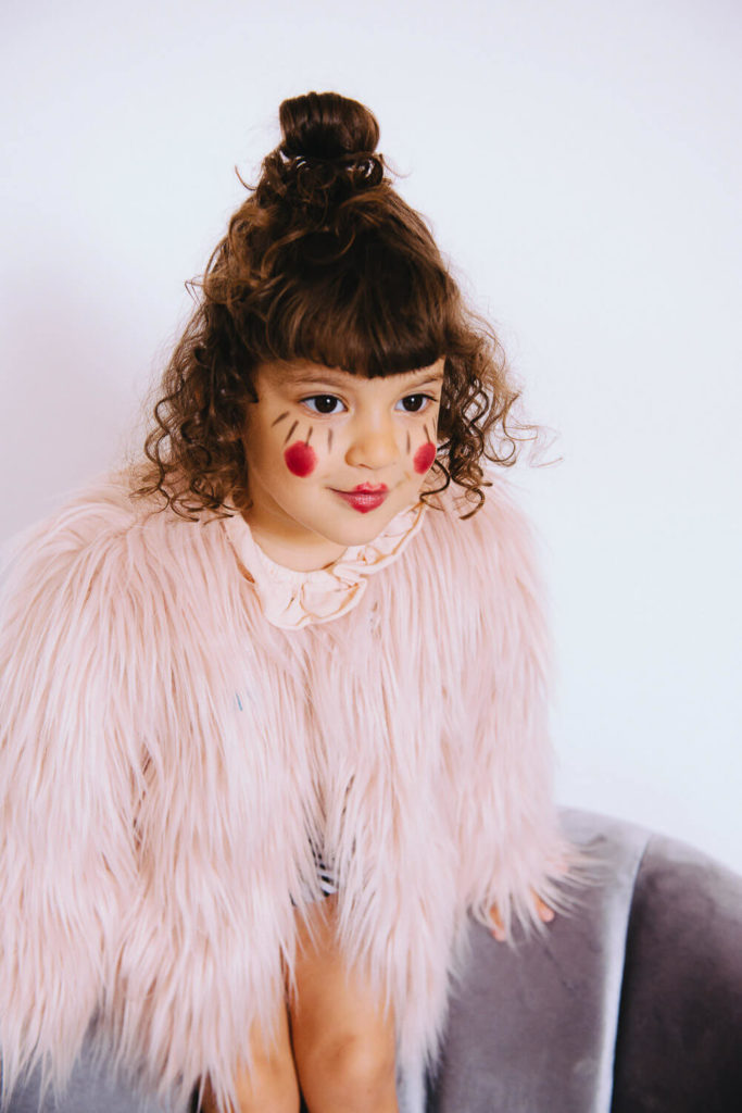 A young child with curly hair and pink cheek paint sits on a gray chair. Theyre wearing a fluffy pink coat and have a topknot hairstyle, looking ahead with a focused expression.