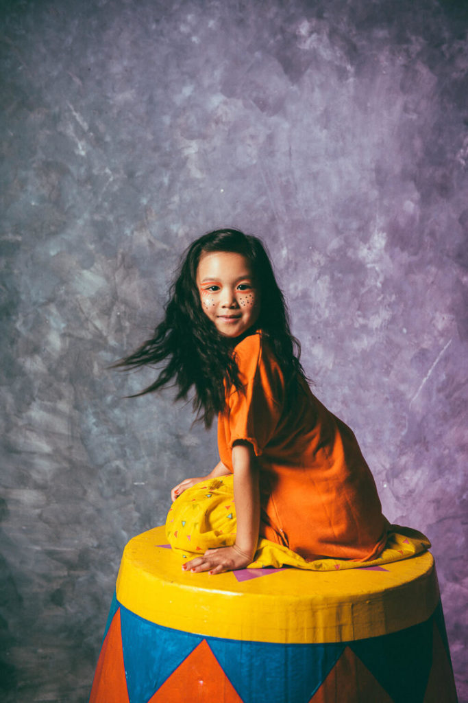 A young girl with painted cheeks smiles while sitting on a colorful, circus-themed platform. She wears an orange outfit and has long, flowing dark hair. The background is a textured purple-gray.