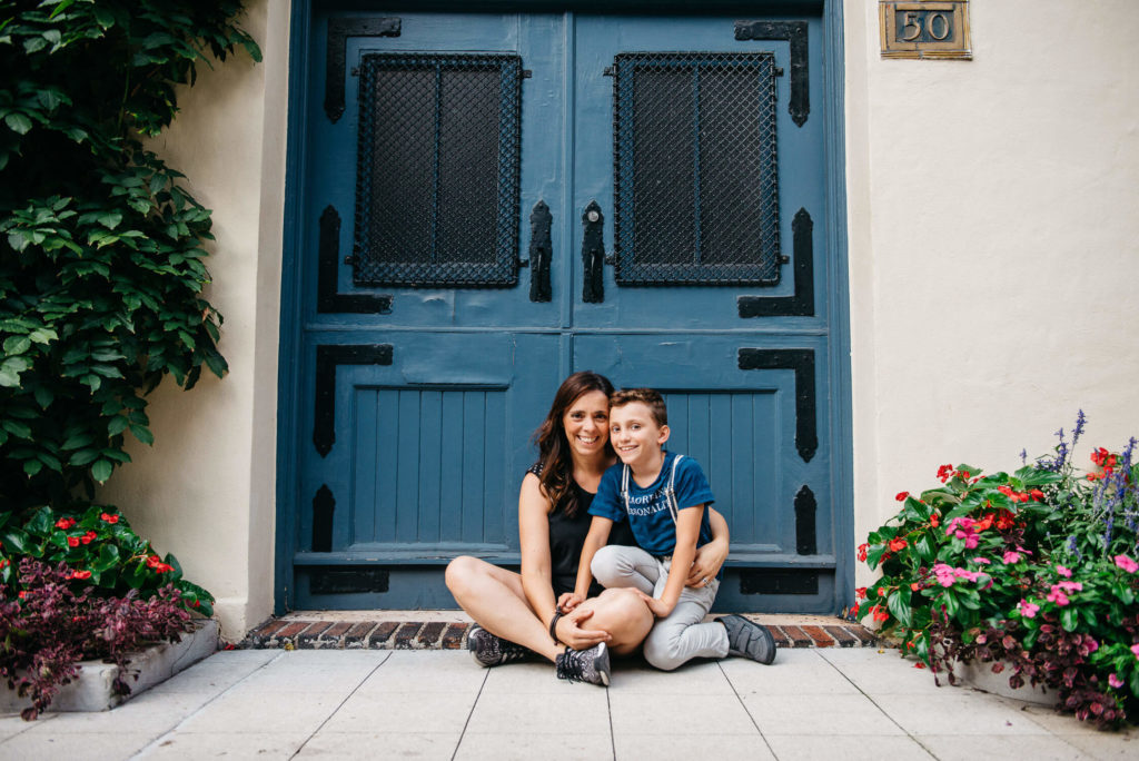 A woman and a child sit on the ground in front of a large blue door. They are surrounded by vibrant flowers and green plants. The woman is smiling and the child is leaning toward her, also smiling.