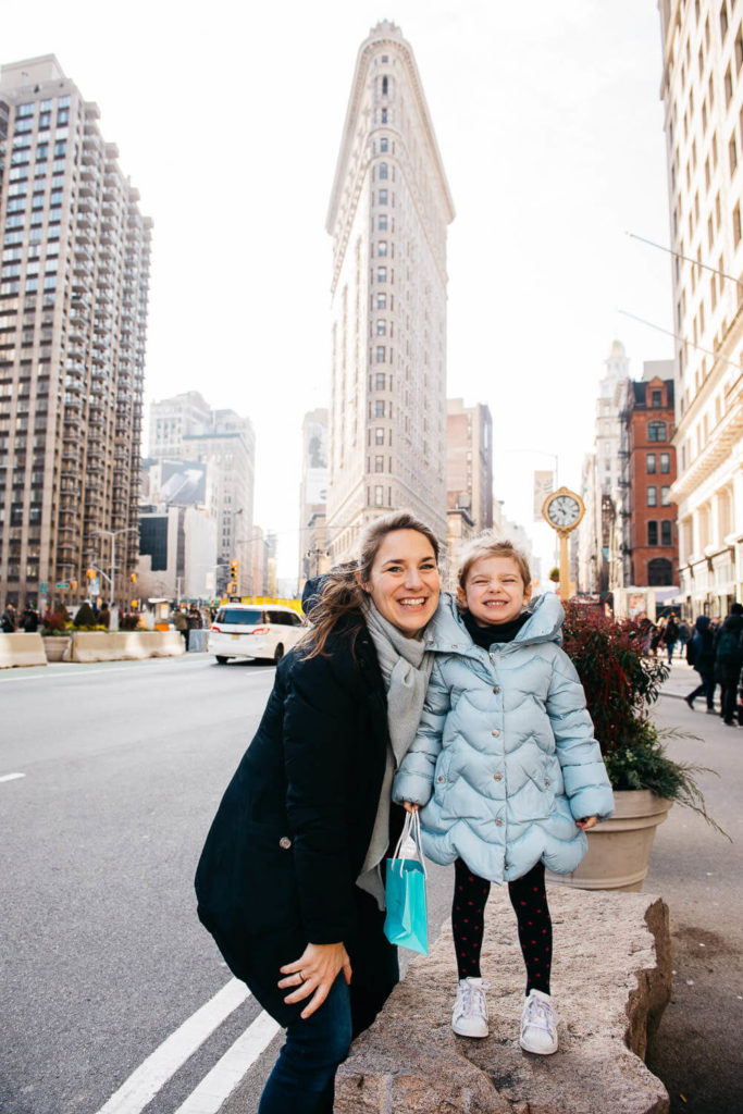 A woman and a child smile at the camera in front of the Flatiron Building. The woman wears a black coat and scarf, and the child is in a light blue coat. Tall city buildings surround them, with a bustling street in the background.