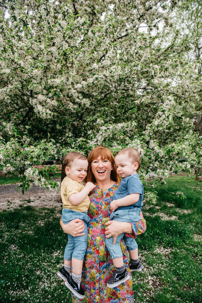 A person joyfully holds two toddlers, one in each arm, in front of a blooming tree. The person is wearing a colorful dress, and the children are dressed in casual clothes. The background features lush greenery and blossoms.