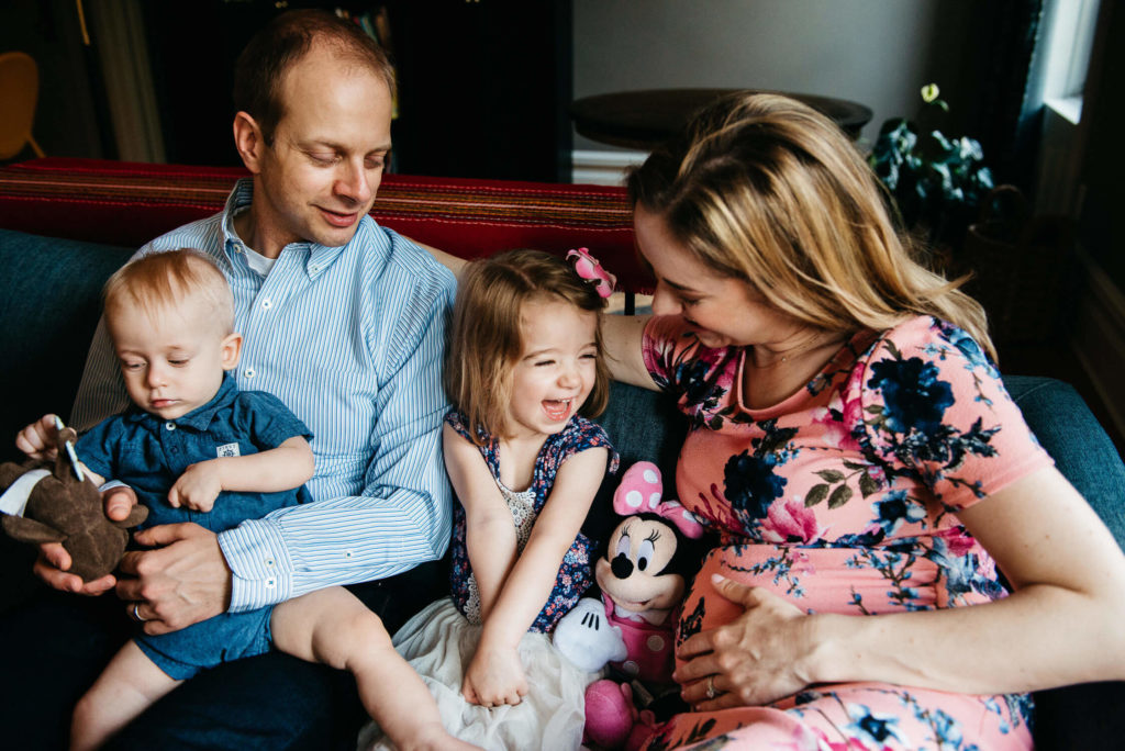 A family sits on a couch. The father holds a baby wearing a blue outfit and a plush toy. The mother, in a floral dress, smiles while holding a little girl with a flower headband and a Minnie Mouse plush toy. The girl is laughing joyfully.