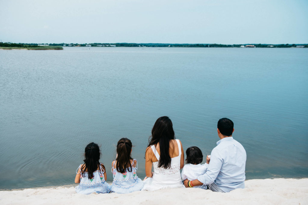 A family of five sits on a sandy beach facing a calm, expansive lake under a clear blue sky. The parents are seated with their three children, all wearing light-colored summer clothing, enjoying a tranquil moment by the water.