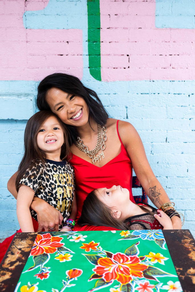 A smiling woman in a red dress sits with two children. One child wears a leopard print top, and the other rests their head on the womans arm. They are in front of a colorful, patterned table and a painted brick wall.