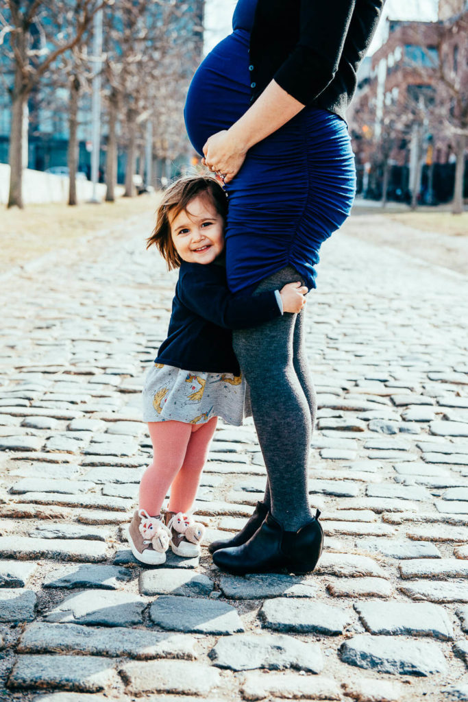 A pregnant woman in a blue dress stands on a cobblestone path, gently holding her belly. A young child with a big smile hugs her leg, wearing a black sweater, gray skirt, pink tights, and brown shoes. Trees and buildings are in the background.