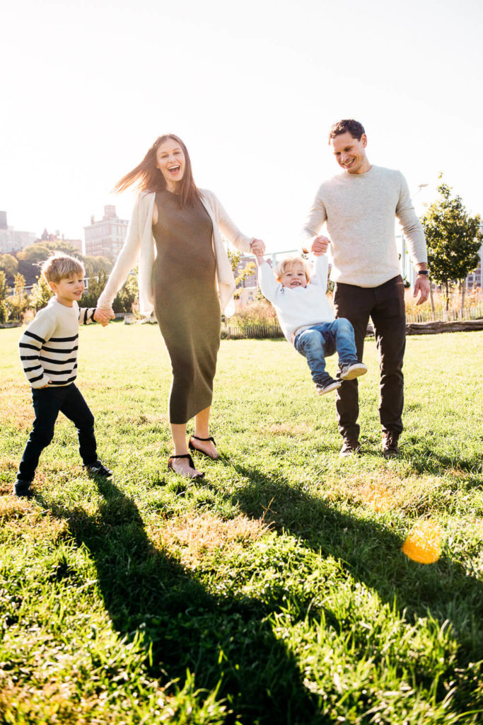 A cheerful family of four enjoys a sunny day in the park. The parents hold their young child by the hands, swinging them in the air, while the older child walks beside with a big smile. The background features a city skyline and green trees.