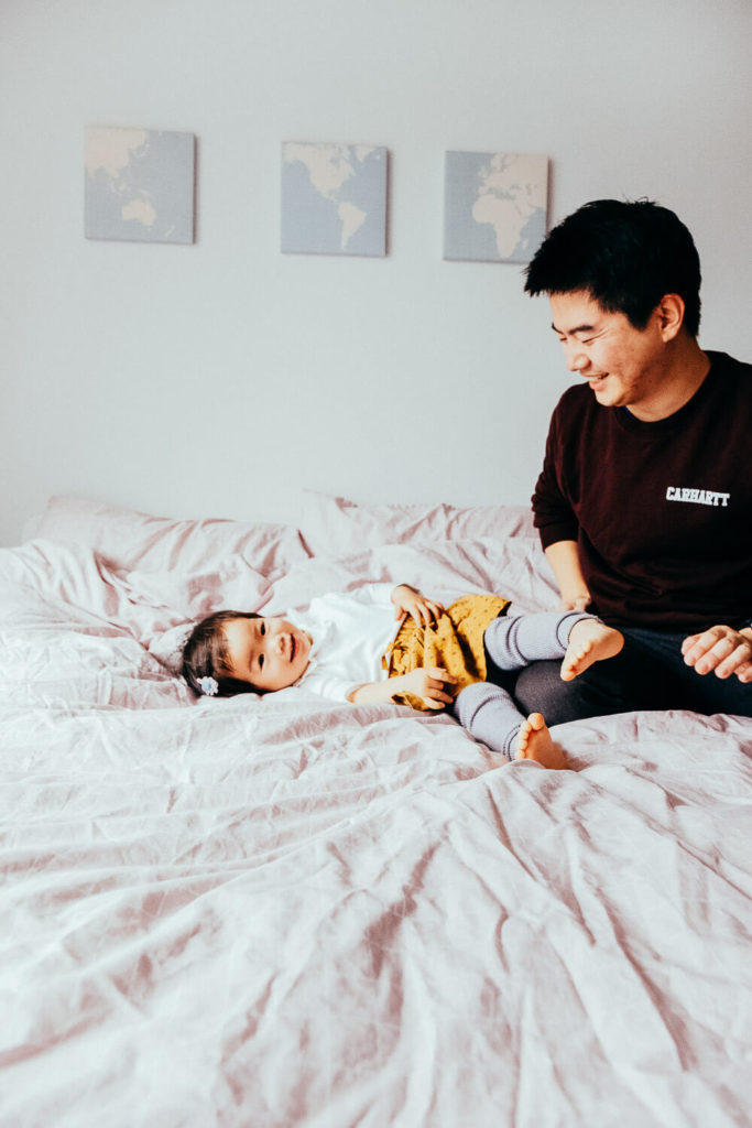 A man and a baby are sitting on a bed. The baby is smiling and lying back, wearing a white shirt and yellow shorts. The man, wearing a dark shirt, is looking at the baby with a smile. Three map prints hang on the wall behind them.