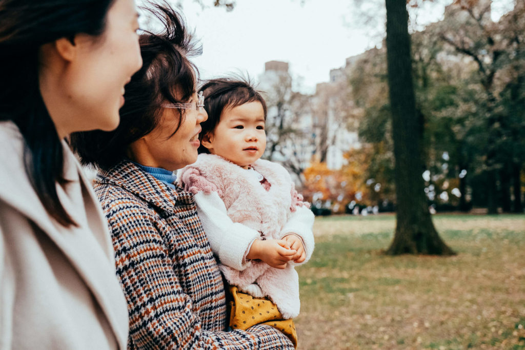 Three people are outdoors in a park. An adult holds a small child, who is wearing a pink and white jacket. The wind is blowing, rustling their hair. Trees and grass are visible in the background.