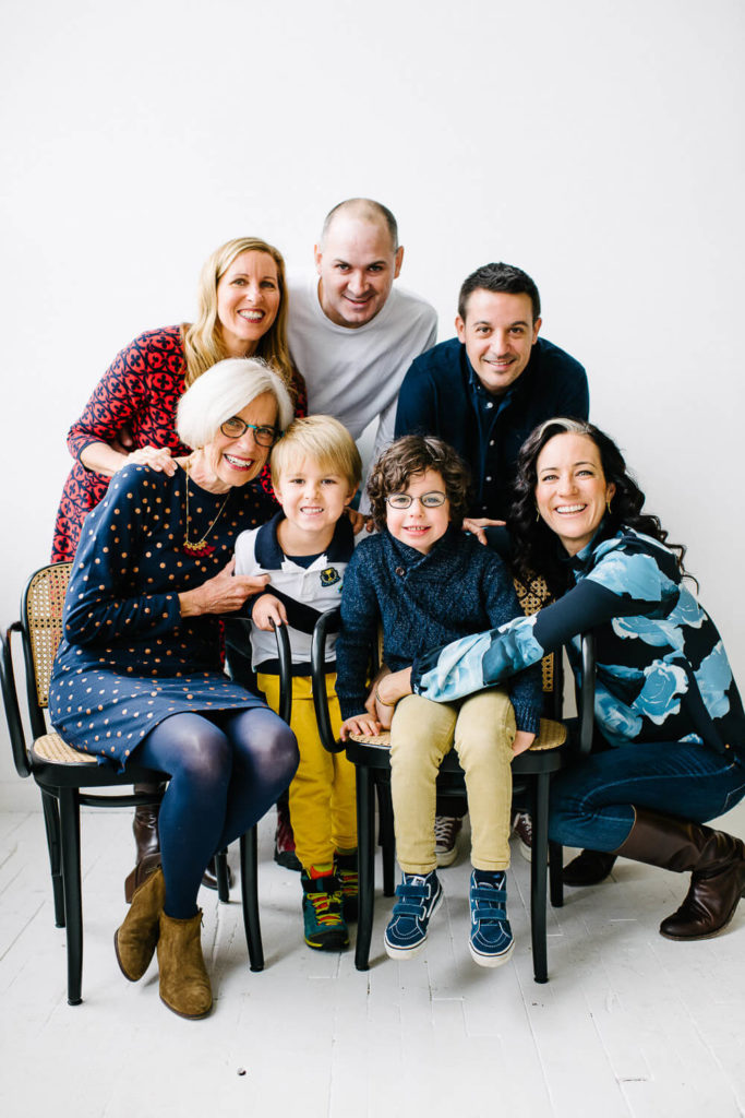 A cheerful family photo with seven people. Two children sit in front, smiling. Behind them are five adults, three women and two men, all smiling and gathered closely. The setting is a bright room with a white background.