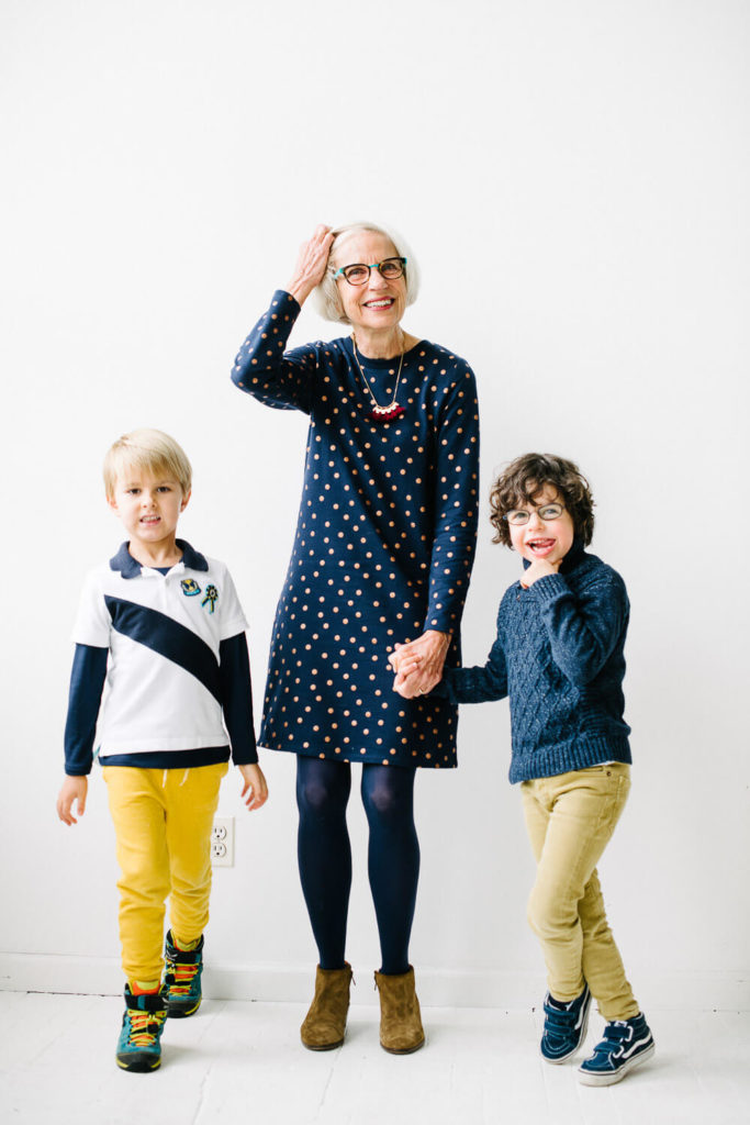 An elderly woman in a navy polka dot dress stands smiling between two young boys. The boy on the left wears a white shirt with a diagonal stripe and yellow pants. The boy on the right wears glasses, a blue sweater, and khaki pants. They are holding hands.