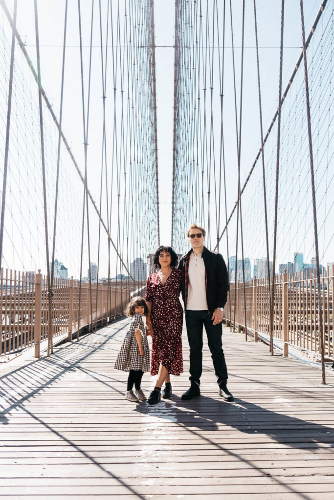 A family of three stands on the Brooklyn Bridge. A woman, man, and young child pose together, with cables and the city skyline visible in the background. The woman wears a red dress, the man a jacket, and the child a plaid dress.